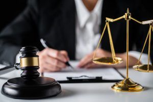 Close-up view of a businesswoman or female lawyer signing a contract with a judge’s gavel and brass scales on the desk, symbolizing law and justice.