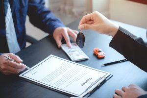 The image representing the concept of a leased car, with a car salesman delivering a toy model car to a new car buyer, while they sign a lease contract in a dealership setting. 