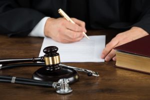 Midsection of a judge writing on legal documents, with a gavel and stethoscope on the desk in a courtroom.