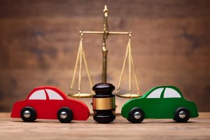 Close-up of two small red and green toy cars placed in front of a justice scale on a wooden desk, symbolizing legal disputes involving car accidents or insurance claims. 