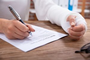 A close-up image of a businesswoman with a white bandaged hand, seated at a wooden desk, filling out a work injury claim form. 