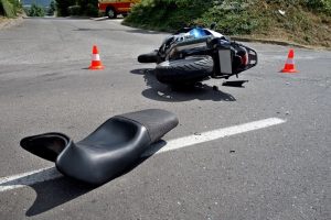 A serious motorcycle accident scene on a sunny day, showing the aftermath of a collision with another vehicle. Close-up view of the damaged motorcycle and debris scattered on the road.