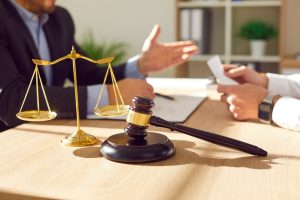 Close-up photo of a male lawyer in a professional office setting, engaged in discussion with a male client across the desk. The desk features legal elements, including a judge's gavel, scales of justice, and neatly arranged documents.